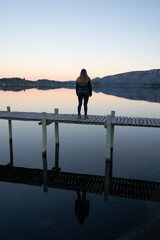 Magical portrait of a young woman standing on the wooden dock in the calm lake at sunset. Its...