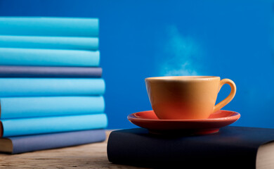 Yellow cup of coffee or tea with hot steam, on book with light blue books and blue background.