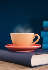 Yellow cup of coffee or tea with hot steam, on book with light blue books and blue background.