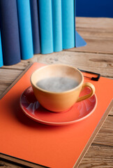 Yellow cup of coffee or tea with hot steam, on note book, with light blue books and blue background.