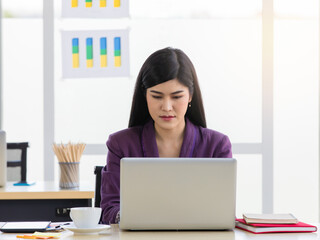 Millennial Asian female businesswoman in formal outfit sitting at work desk with coffee cup tablet post it and notebook using laptop computer browsing internet website check email in company office