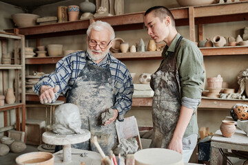 Senior man in apron holding the lesson for woman in pottery workshop, he teaching her to make...