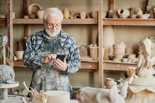 Senior sculptor in apron typing message on mobile phone while working in his pottery studio