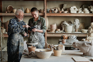 Senior sculptor pointing at tablet pc and showing his assistant the ceramic sculpture during their work in pottery studio - obrazy, fototapety, plakaty