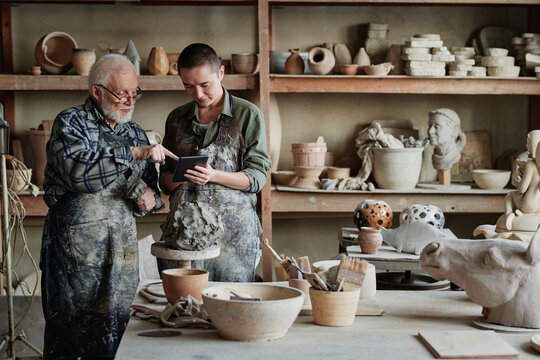 Senior sculptor pointing at tablet pc and showing his assistant the ceramic sculpture during their work in pottery studio