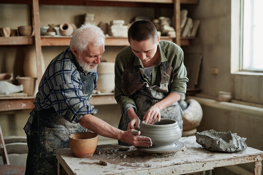 Young female potter making earthenware vase on pottery wheel with senior man helping her during their work in workshop