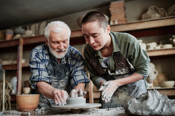 Senior man showing the woman how to make form of vase from clay on pottery wheel in studio - obrazy, fototapety, plakaty