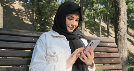 Woman in black hijab using smartphone in city park, close-up. Female siting on bench looking at phone. Young muslim woman in traditional black head scarf smiling holding smart phone on sunny street