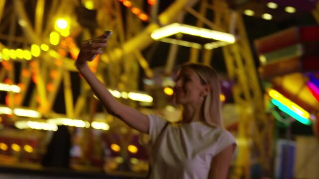 Young blonde woman enjoys bright lights of carnival while taking a selfie. Handheld shot.