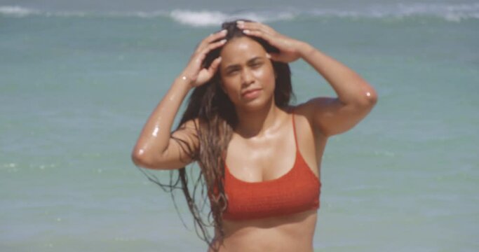 Medium shot of young beautiful Latin American girl wearing red bikini on exotic beach walks towards camera while tightening hair, Dominican Republic, handheld, day