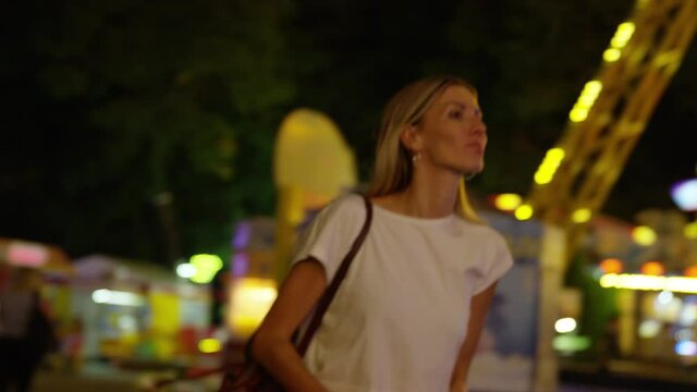 Young blonde woman turns around and marvels at carnival lights before taking photo. Tracking shot.