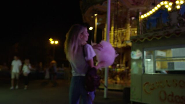 Young blonde woman enjoys carnival at night time and eats candy floss. Tracking shot.