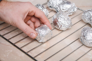 The man wraps peeled raw potatoes in foil and places them on the wire rack to bake them in the oven.