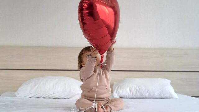 little Caucasian girl sitting on the bed holding a red heart-shaped balloon with helium inside. concept of celebrating valentine's day on february 14.