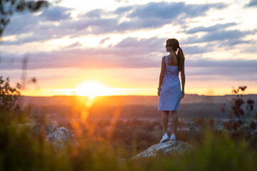 Dark silhouette of a young woman standing on a stone enjoying sunset view outdoors in summer