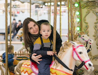 Mom and son are having fun on the merry-go-rounds. Child boy rides a carousel horse, amusement.