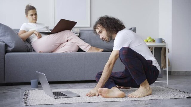 Curly Guy Doing Yoga Exercise Before Laptop On Floor And Lady With Notebook Relaxing On Sofa At Home. Woman Sits On Man During Routine Workout Couple Fall On Carpet And Have Fun