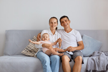 Indoor shot of smiling happy couple sitting on cough with toddler daughter in living room, looking...