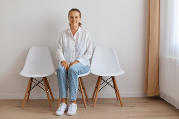 Fototapeta premium Indoor shot of attractive young adult woman sitting on chair against white wall, wearing shirt and jeans, posing with smile, looking happily at camera, expressing optimism.