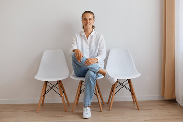 Horizontal shot of cheerful attractive young adult woman sitting on chair against white wall, wearing shirt and jeans, sincerely smiling at camera, positive mood.