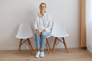 Fototapeta premium Image of smiling satisfied attractive young adult woman sitting on chair against white wall, wearing shirt and jeans, looking at camera with calm and optimistic facial expression.