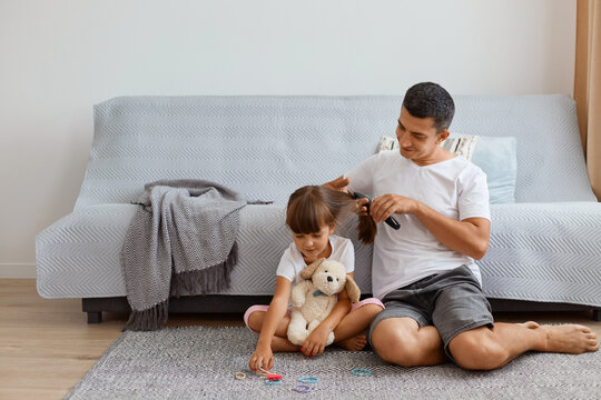 Positive happy man with dark hair wearing white t shirt combing daughter's hair while sitting on floor near cough, making ponytail for her kid before going to school or kindergarten.