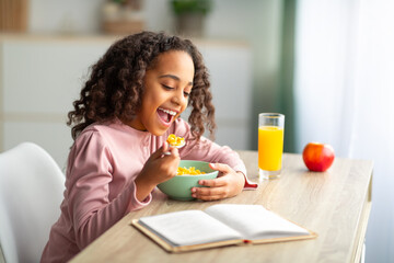 Cute black teen girl having healthy breakfast and reading book while sitting at desk at home