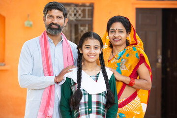 Portrait of happy Indian family standing outside their house in village Smiling Parents with their adorable young daughter in traditional outfit. rural india concept. looking at camera