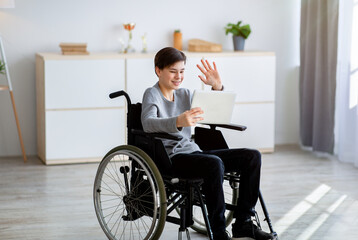 Positive handicapped teenager in wheelchair having video conference with teacher on tablet pc, waving at screen indoors
