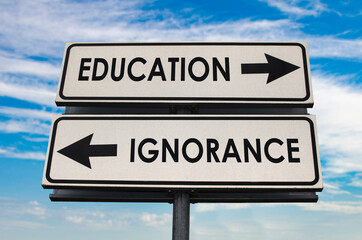 Education versus ignorance road sign with two arrows on blue and grey sky background. White two street sign with arrows on metal pole. Two way road sign with text. - obrazy, fototapety, plakaty