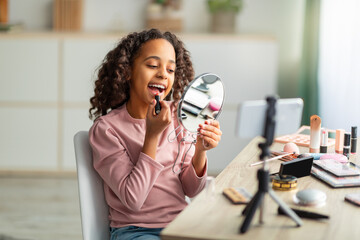 Beautiful african american teen blogger applying lipstick and looking at mirror while filming vlog for her channel