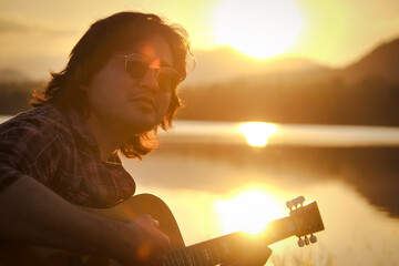 Man playing acoustic guitar outdoors with sunlight reflected on water surface at sunset lake.