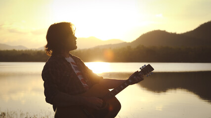 Man playing acoustic guitar outdoors with sunlight reflected on water surface at sunset lake.