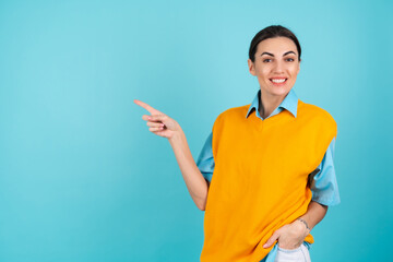 Young woman in a shirt and an orange vest on a turquoise background smiling cheerfully, pointing her finger to the left to an empty space