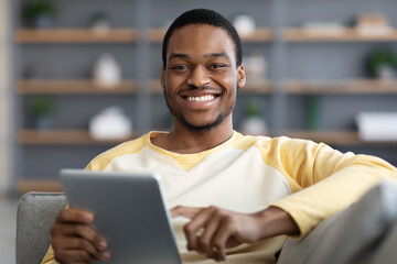 Joyful black guy resting with digital tablet on couch