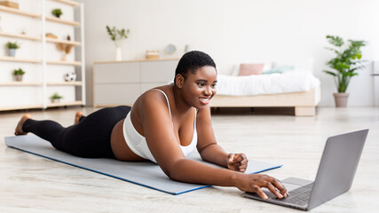 Overweight black woman lying on sports mat, using laptop, communicating with personal coach or watching fitness video