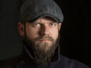 portrait of a bearded young man in a vintage cap close-up, looking into the camera. the collar of his coat is turned up