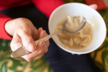 top view of hand of old woman holding tablespoon of chicken noodle soup (focus on the upper foreground thumb)
