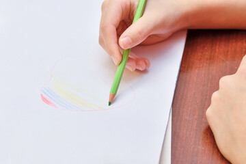 The girl draws a heart on a white sheet of paper and paints it in the form of a rainbow with colored pencils.