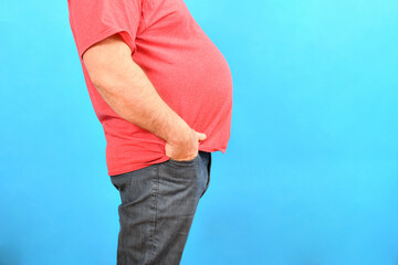 A fat man with a big belly in jeans and a red T-shirt on a blue background holds his hands in his...