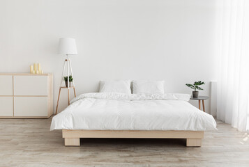 Real photo of bedroom interior in simple style. Big bed with pillows, lamp and furniture, white wall, empty space