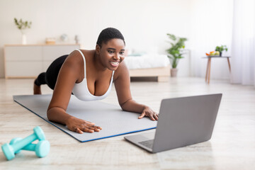 Plump black lady exercising at home, following personal trainer's online instructions on laptop,...
