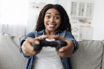 Weekend leisure. Overjoyed african american woman using joystick and playing video games, sitting on sofa at home © Prostock-studio