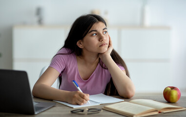 Thoughtful Indian teen girl studying online from home, taking notes during web lesson, feeling...