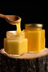 on a wooden log there are two glass jars with honey on a dark background. take honey out of the jar with a wooden spoon.