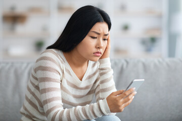 Worried Asian Lady Looking At Smartphone While Sitting On Sofa At Home