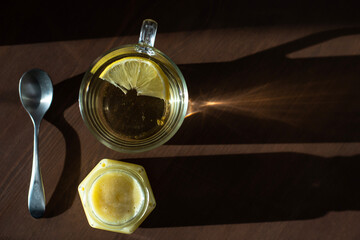 top view of a mug of tea with lemon, a spoon and a jar of creamy honey on a wooden table. sunbeams are reflected from a glass mug on the table.