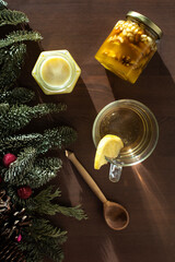 top view of tea with lemon in a cup, decorated with Christmas tree twigs and two jars of honey with a spoon on a wooden table top