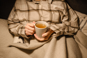 A cup of tea in the hands of a girl. The girl is drinking hot tea. A girl in pajamas is wrapped in a blanket and is drinking delicious tea. Enjoy the comforts of home. White cup close up in woman hand