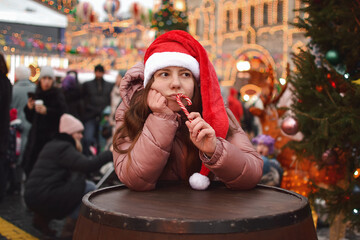 A young bored woman wearing a Santa hat at the Christmas market is sad, leaning on her hand and looking away, holding a cane-shaped candy bought at the market in her hand and pressing it to her lips.